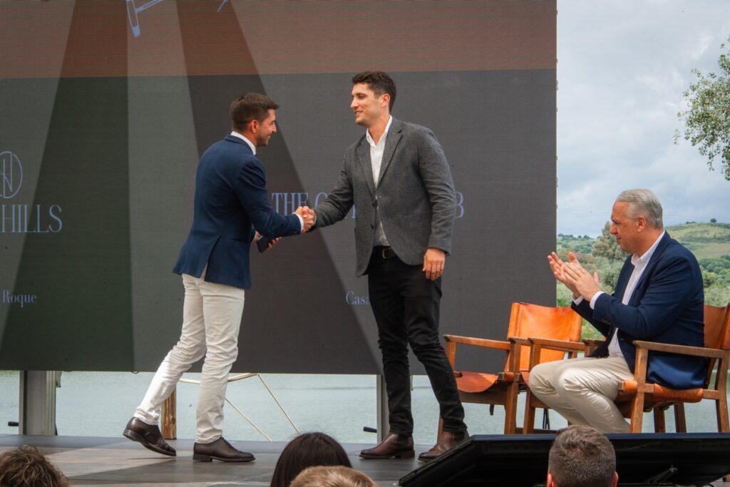 Two men shake hands on stage at a Sotogrande investment event, while a third man applauds from his seat in front of a scenic lake backdrop.