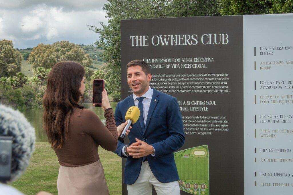A man in a blue suit speaks to a reporter in front of a sign for The Owners Club during a Sotogrande investment event by Polo Valley.