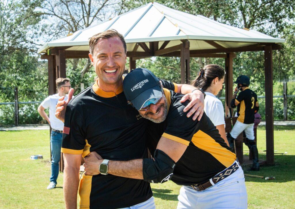 Two smiling men in black and yellow polo uniforms share a joyful moment during a polo event in Sotogrande, with teammates and a wooden gazebo in the background.