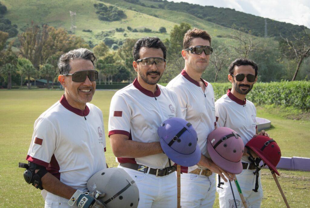 Four polo players in matching white and maroon uniforms pose with their helmets at a lush green field in Sotogrande.