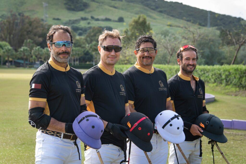 Four male polo players in black and gold uniforms hold colorful helmets while standing on a lush polo field in Sotogrande.