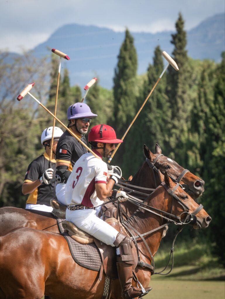 Polo players on horseback in mid-match at the Sotogrande polo grounds, holding mallets against a scenic mountain and forest backdrop.